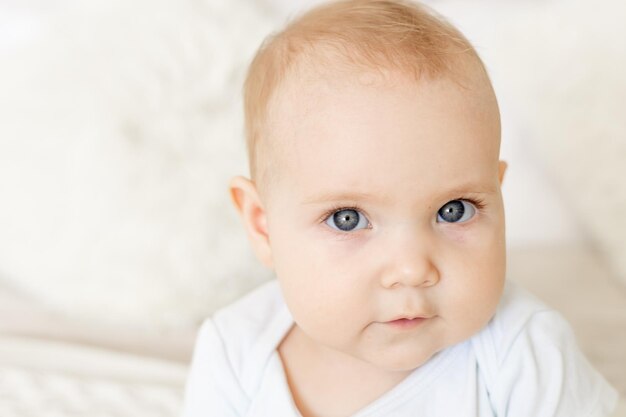 Closeup do retrato de um menino ou menina de seis meses sentado em casa em uma cama em um quarto brilhante e sorrindo ou rindo de um recém-nascido feliz em uma roupa branca com olhos azuis