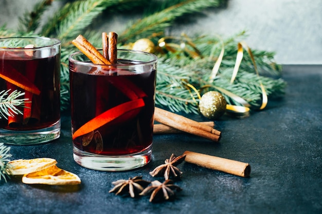 Closeup em uma bebida alcoólica quente vinho quente em dois copos transparentes com um pau de canela laranja e anis estrelado em uma mesa escura no contexto de galhos de árvores de Natal