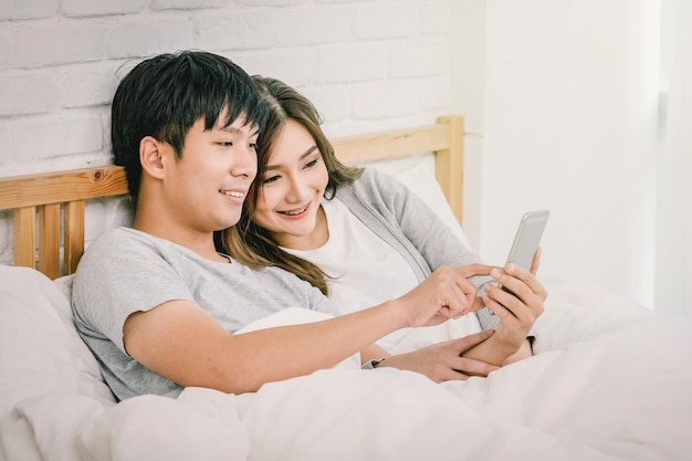 Closeup feliz amante asiático o pareja usando tecnología de teléfonos inteligentes en la cama