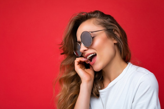 Closeup foto de encantadora mujer rizada rubia oscura joven feliz aislada sobre pared de fondo rojo con camiseta blanca casual y gafas de sol con estilo mirando hacia el lado.