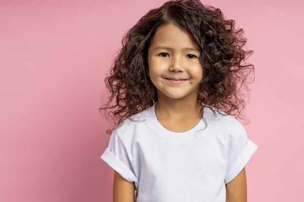 Closeup foto de encantadora niña caucásica en camiseta blanca, con mirada amable y amable mirando con linda sonrisa, posando contra la pared rosa. Infancia feliz, inocencia infantil, concepto de niños