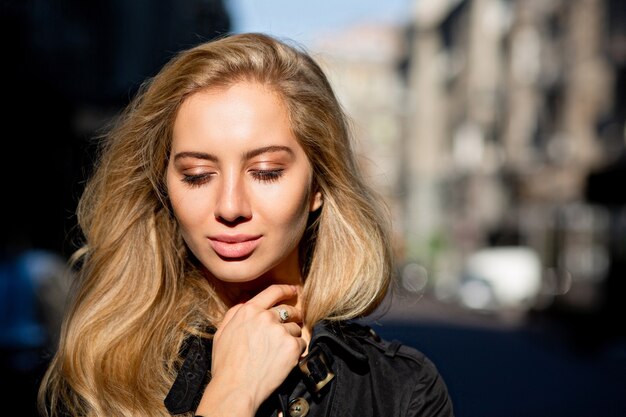 Closeup foto de increíble mujer rubia con cabello largo posando en el fondo de los edificios de la ciudad. Espacio para texto