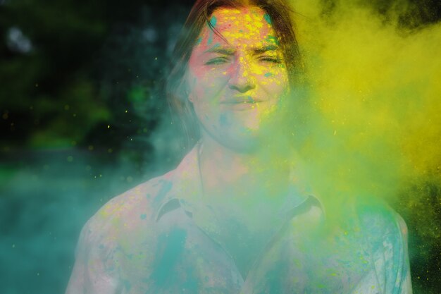 Closeup foto de mujer atractiva con pelo corto posando con pintura Holi amarilla y verde explosiva en el parque. Espacio para texto