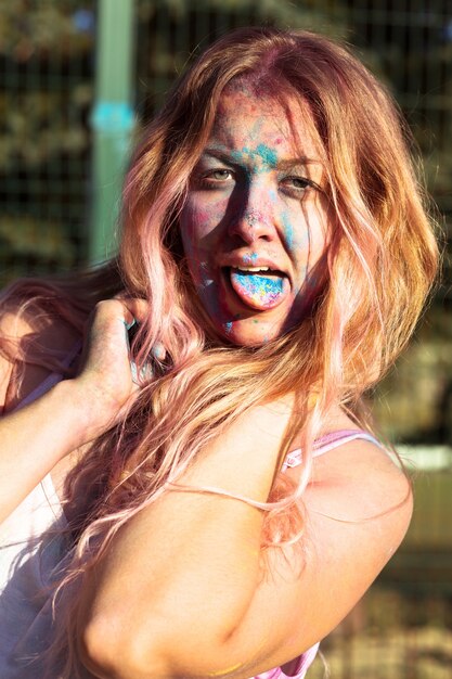 Closeup foto de mujer rubia expresiva cubierta con pintura colorida Holi, mostrando su lengua