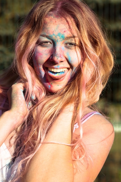 Closeup foto de mujer rubia positiva cubierta con pintura colorida Holi, mostrando su lengua