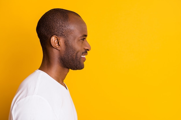 Closeup foto de perfil de atractivo alegre chico de piel oscura buen humor radiante sonrisa mirada espacio vacío usar camiseta blanca casual aislado fondo de color amarillo vivo brillante