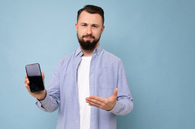 Closeup foto de triste insatisfecho guapo guapo joven morena sin afeitar con barba vistiendo camiseta blanca casual y camisa azul poising aislado sobre fondo azul con espacio vacío sosteniendo