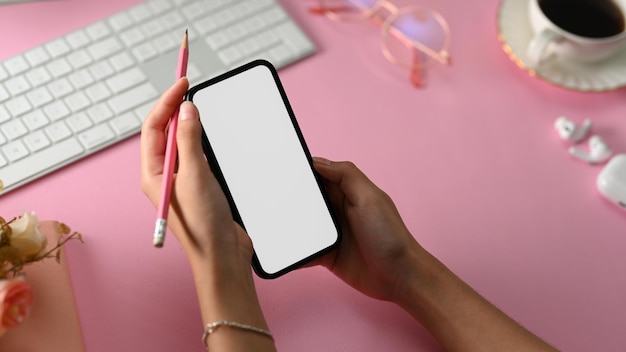 Closeup Girl sosteniendo una maqueta de pantalla en blanco de teléfono celular y un lápiz sobre su elegante escritorio de oficina rosa