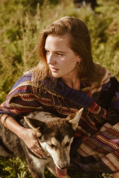 Foto closeup hermosa chica sin maquillaje en una capa etno se sienta en la hierba del prado y acaricia al perro