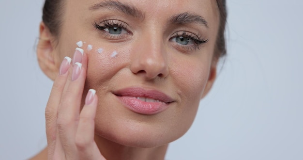 Closeup hermosa mujer joven aplica crema para el cuidado de la cara en la mejilla sonriendo para la cámara sobre fondo gris Concepto comercial de cuidado de la piel