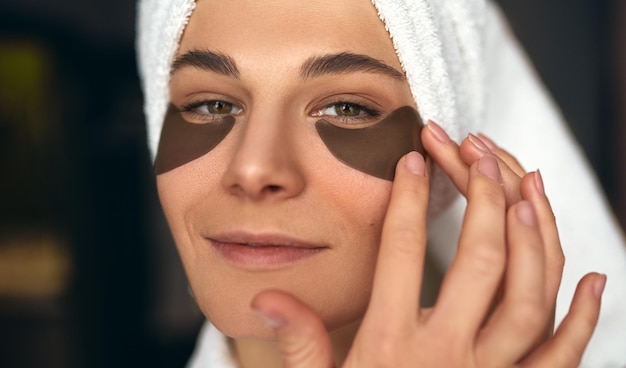 Closeup iamge de mulher bonita depois do banho com toalha na cabeça coloca manchas sob os olhos de rugas e olheiras mulher atraente sorrindo e fazendo tratamentos de beleza no rosto em casa