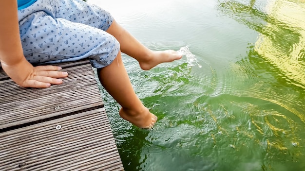 Closeup imagem de criança sentada no cais de madeira em tiver e segurando os pés na água. Crianças brincando e jogando água com as pernas