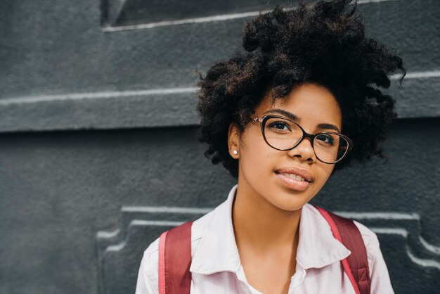 Closeup imagem de jovem estudante americano africano usando óculos se preparando para ir para a faculdade Bela jovem africana sorrindo com mochila posando na parede cinza Educação de pessoas