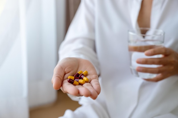 Closeup imagem de uma mulher doente segurando pílulas e um copo de água enquanto está sentado em uma cama