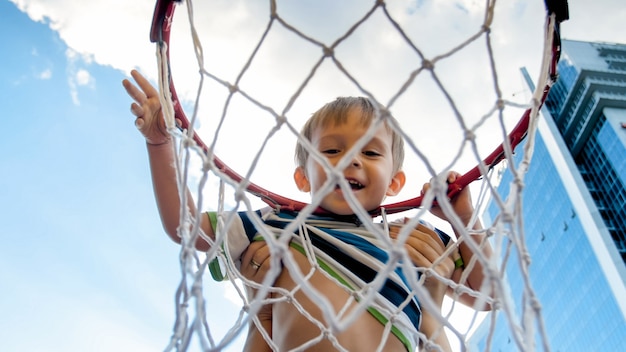 Closeup imagen de lindo niño pequeño sonriente sosteniendo y colgando del aro de baloncesto en el patio de recreo de los deportistas en la ciudad