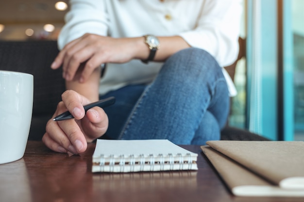 Closeup imagen de la mano de una mujer sosteniendo un bolígrafo para escribir en un cuaderno en blanco con una taza de café en la mesa de madera