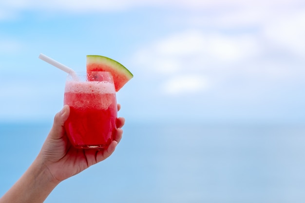 Closeup imagen de una mano sosteniendo un vaso de batido de sandía junto al mar con fondo de cielo azul