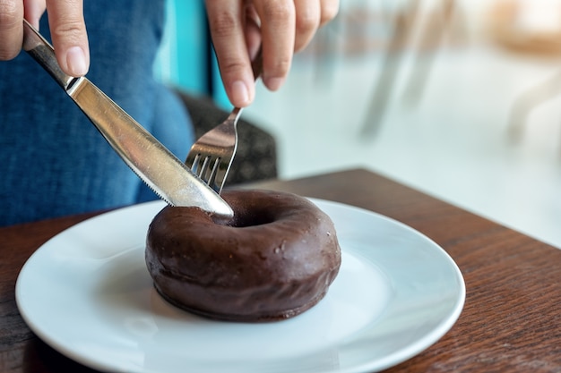 Closeup imagen de manos de mujer cortando un trozo de donut de chocolate con cuchillo y tenedor para desayunar en la mesa de madera