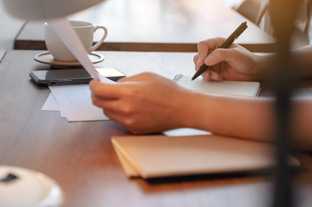 Closeup imagen de manos de mujer sosteniendo papel de documento y escribiendo en un cuaderno en blanco sobre la mesa en el café