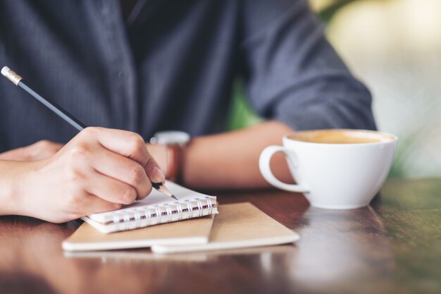 Closeup imagen de una mujer escribiendo en un cuaderno en blanco con una taza de café en la mesa de café