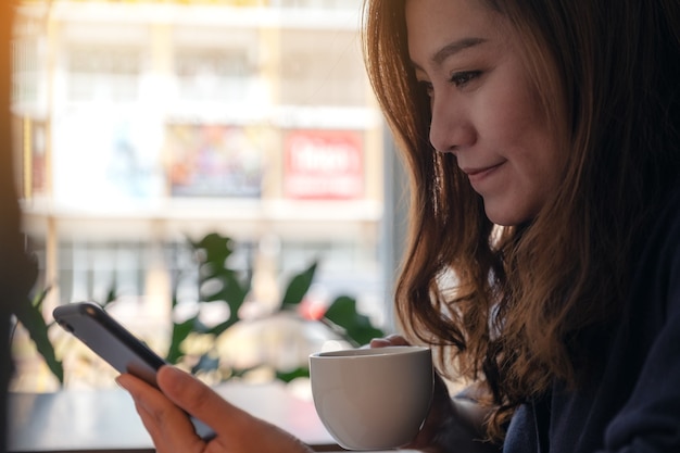 Closeup imagen de una mujer sosteniendo, usando y mirando el teléfono inteligente mientras bebe café en la cafetería