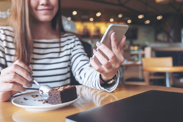 Closeup imagen de una mujer sosteniendo, usando y mirando el teléfono inteligente mientras come pastel de brownie en café