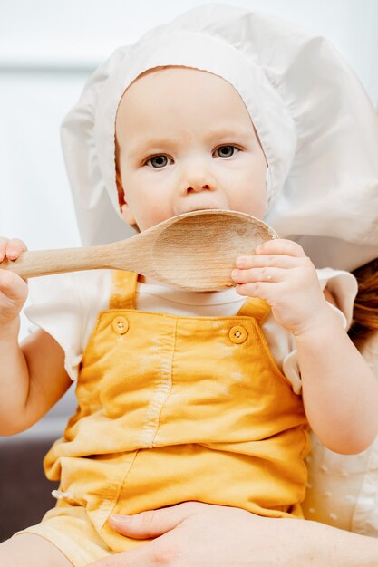 Closeup linda niña pequeña en un traje de cocinero lame una espátula de cocina de madera