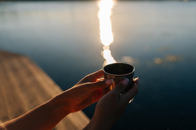 Closeup mãos de mulher irreconhecível segurando xícara com café quente de garrafa térmica sentado na praia pelo cais de madeira na manhã ensolarada de verão