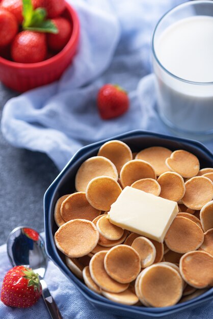 Closeup minúsculas panquecas de cereal com uma fatia de manteiga, morangos e copo de leite no azul