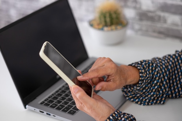 Closeup nas mãos da mulher usando o telefone celular enquanto trabalhava com o site de navegação feminina madura do laptop no smartphone