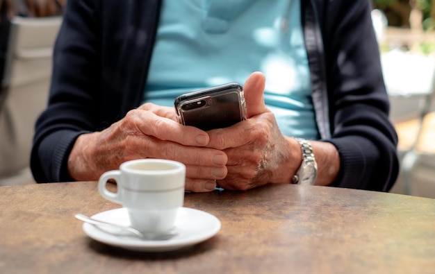 Closeup nas mãos do velho idoso usando telefone celular Homem caucasiano idoso sentado na mesa de café com uma xícara de café expresso enquanto olha para seu smartphone
