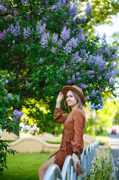 Closeup niña de pie sobre un fondo de arbustos lilas. ella es de apariencia europea con un sombrero y vestido marrones