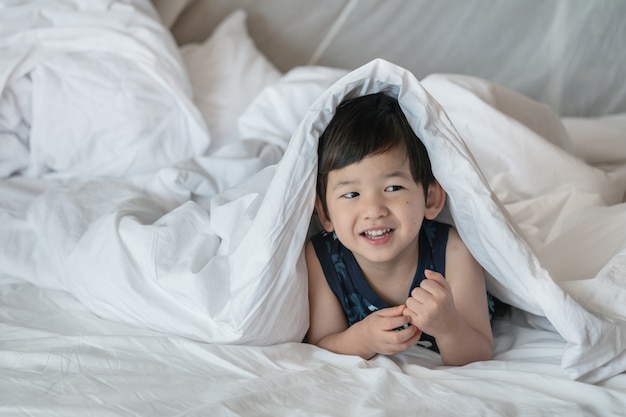 Closeup niño asiático en la cama debajo de la manta con cara de sonrisa en la mañana