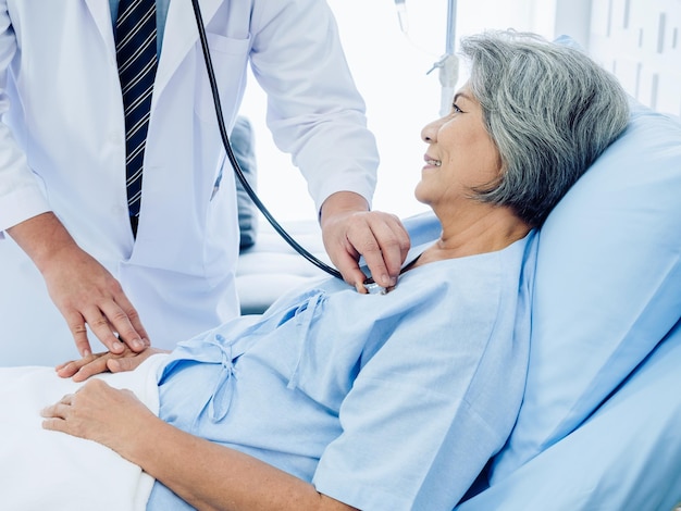 Closeup paciente idosa asiática vestida de azul claro sorrindo alegremente na cama enquanto o gentil médico masculino de terno branco usa estetoscópio para ouvir batimentos cardíacos para check-up no hospital