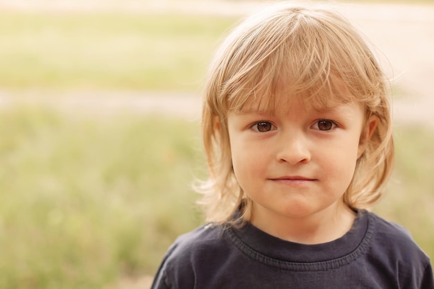 Closeup Portrait des nachdenklichen Gesichts eines kleinen blonden Jungen auf einer Sommerstraße mit grünem Hintergrund