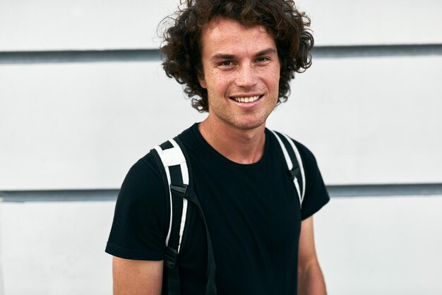 Closeup retrato ao ar livre de homem sorridente bonito com cabelo encaracolado com mochila em pé na construção de parede na rua enquanto espera seus colegas Empresário vestindo camiseta preta casual Pessoas