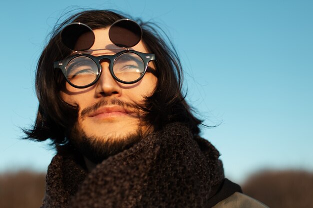 Closeup retrato de chico joven con pelo largo con bufanda y gafas de sol redondas hipster. Cielo azul de fondo.