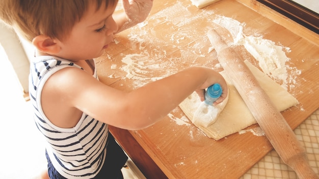 Closeup retrato de adorável menino de 3 anos de idade, fazendo biscoitos e rolando massa com pino de rooling de madeira. pequeno chef cozinheiro