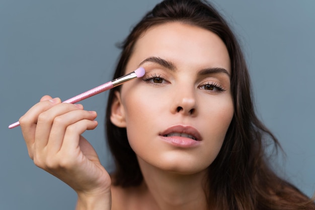 Closeup retrato de beleza de mulher com pele perfeita e maquiagem natural lábios nus cheios segurando um conceito cosmético de pincel de sombra rosa brilhante