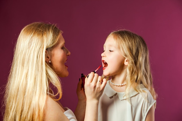 Closeup retrato de encantadora mãe loira e filha no estúdio Mulher colorindo os lábios das meninas pelo batom vermelho líquido brilhante A menina pré-escolar quer ser como sua mãe