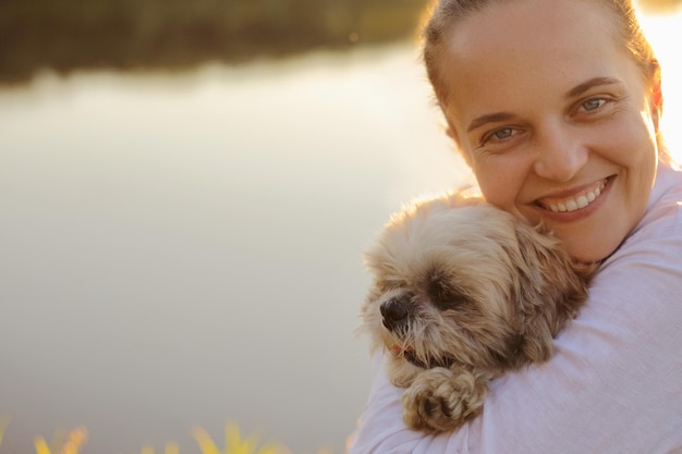 Closeup retrato de feliz satisfeito encantador jovem adulto vestindo camisa branca e seu cachorro pequinês sentado ao pôr do sol perto do rio abraçando seu animal de estimação e olhando para a câmera