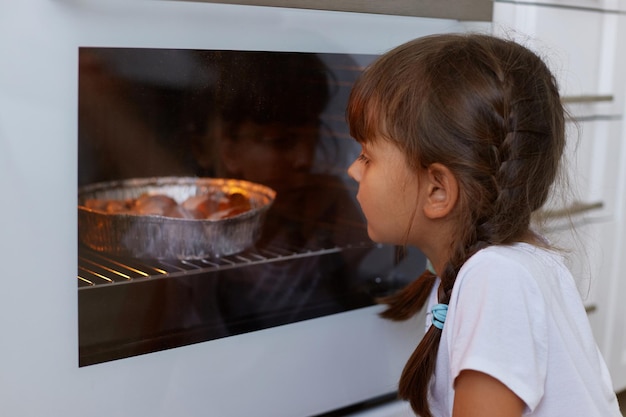 Closeup retrato de menina de cabelos escuros com tranças sentado perto do forno no chão na cozinha e olhando para dentro esperando saborosos bolinhos ou biscoitos vestindo camiseta branca