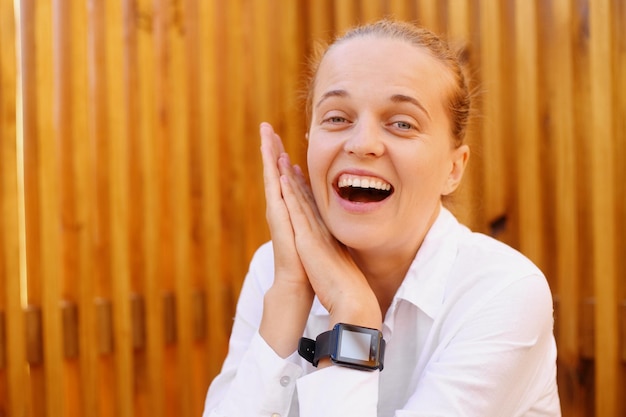 Closeup retrato de mulher rindo extremamente feliz com penteado de coque vestindo camisa branca sentado com as palmas das mãos juntos sendo muito satisfeito olhando para a câmera gritando alegremente