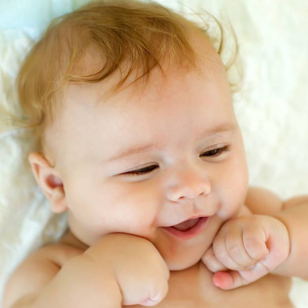 Closeup retrato de um lindo sorridente garotinho engraçado com cabelo loiro deitado na cama com um cobertor macio olhando para longe, interior, imagem quadrada