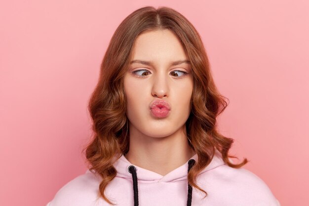 Closeup retrato de uma adolescente engraçada com cabelo encaracolado, fazendo cara de boba com olhos cruzados, expressão estúpida desajeitada se divertindo tiro de estúdio interior isolado no fundo rosa