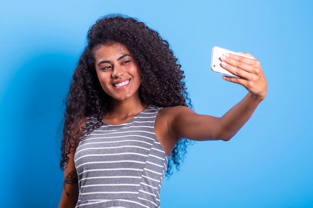 Closeup retrato de uma jovem e atraente mulher afro-brasileira sorridente, segurando um smartphone, tirando uma foto de selfie no fundo azul