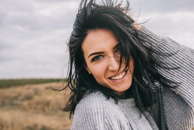 Closeup retrato de uma jovem morena feliz vestindo suéter cinza sendo brincalhão e despreocupado com lindo sorriso e cabelos ventosos posando na paisagem Conceito de estilo de vida Cobrir o humor da ideia