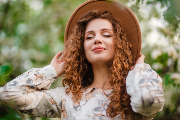 Closeup retrato de uma jovem mulher aproveitando o dia ensolarado de primavera no jardim florido. Mulher com cabelo castanho encaracolado, vestido branco e chapéu marrom entre árvores floridas.