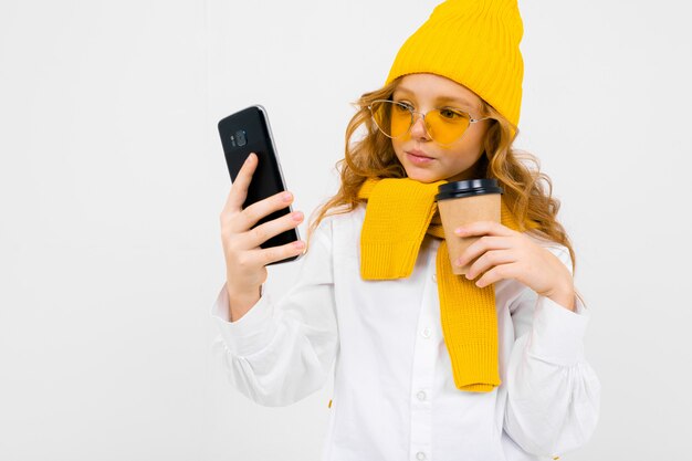 Closeup retrato de uma menina adolescente bonito caucasiano em um inverno chapéu e cachecol com um telefone na mão e um copo de papel tomando uma selfie em branco