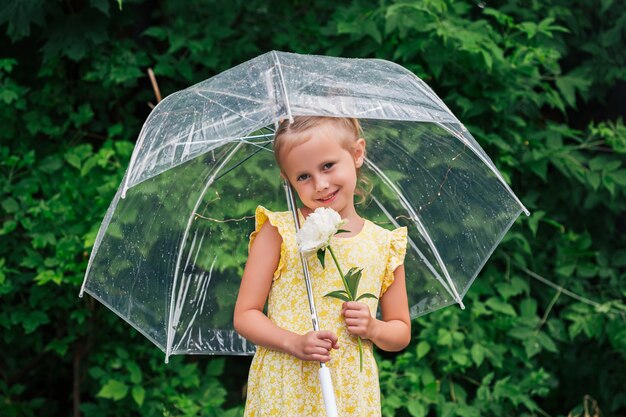 Closeup retrato de uma menina caucasiana com guarda-chuva transparente em um vestido amarelo e botas de chuva no parque durante o verão